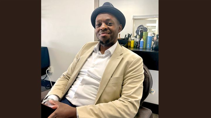 A middle-aged man in a hat, white shirt, and tan jacket relaxes in a chair in a barber shop.