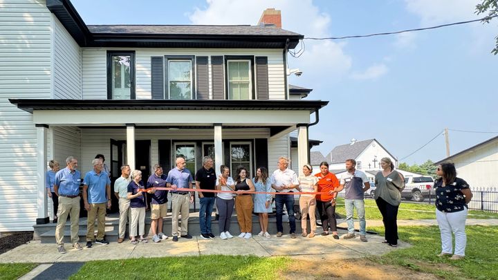Several people stand in a row behind a large ribbon in front of a two-story white house with brown trim.