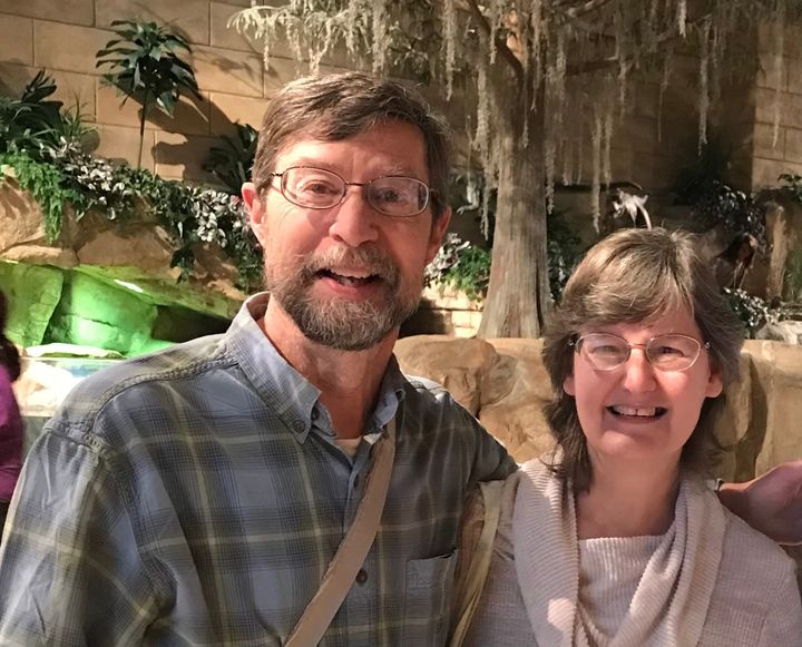 A smiling man and woman pose together indoors in front of a nature-themed backdrop with rocks, plants, and trees.
