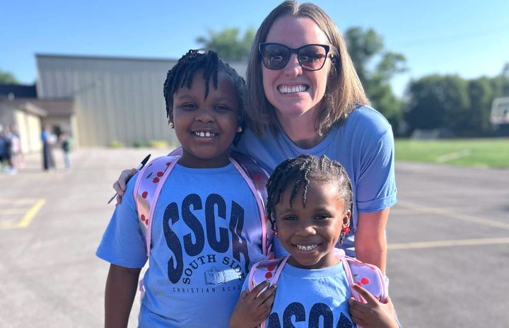 Two smiling children and a woman wearing "South Side Christian Academy" shirts and backpacks outside.