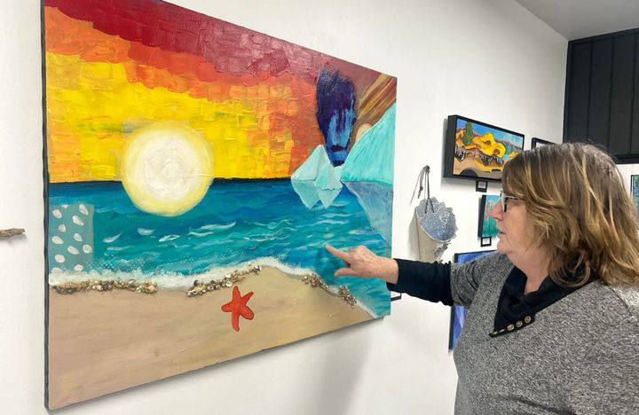 A woman in a black and gray outfit points to a mixed-media work in an art gallery.