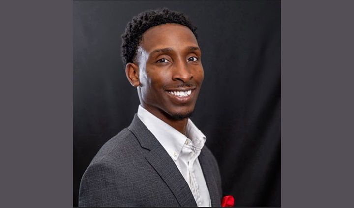 A young man wearing a white shirt and gray blazer smiles at the camera.