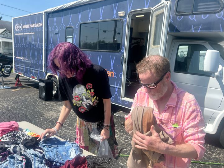 A woman holding a bag and a man holding clothing look at tables full of clothes. The woman is wearing a black shirt with a design on it. The man has a pink plaid shirt and sunglasses.