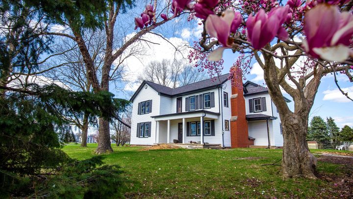 A two-story white house framed by trees stands under a blue sky with bright white clouds.
