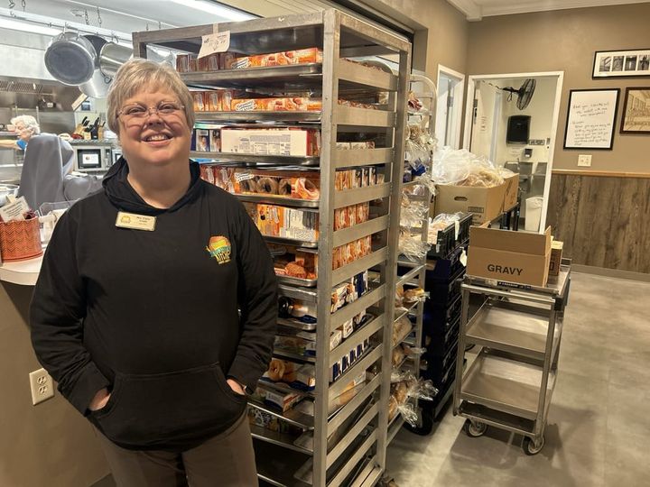 A woman in a brown hoodie stands in front of a rack of pastries in a busy kitchen.