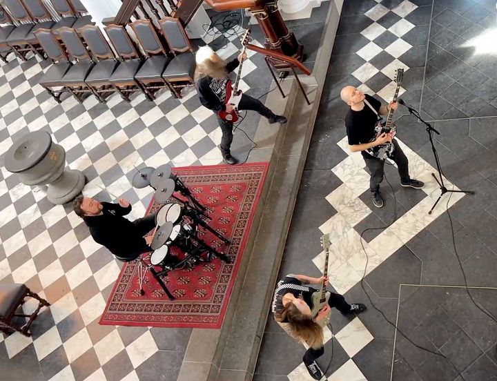 Overhead view of Christian metal band At the Mountain performing on a checkered stone floor inside a church, with guitarists, a bassist, and a drummer set up on a red patterned rug.