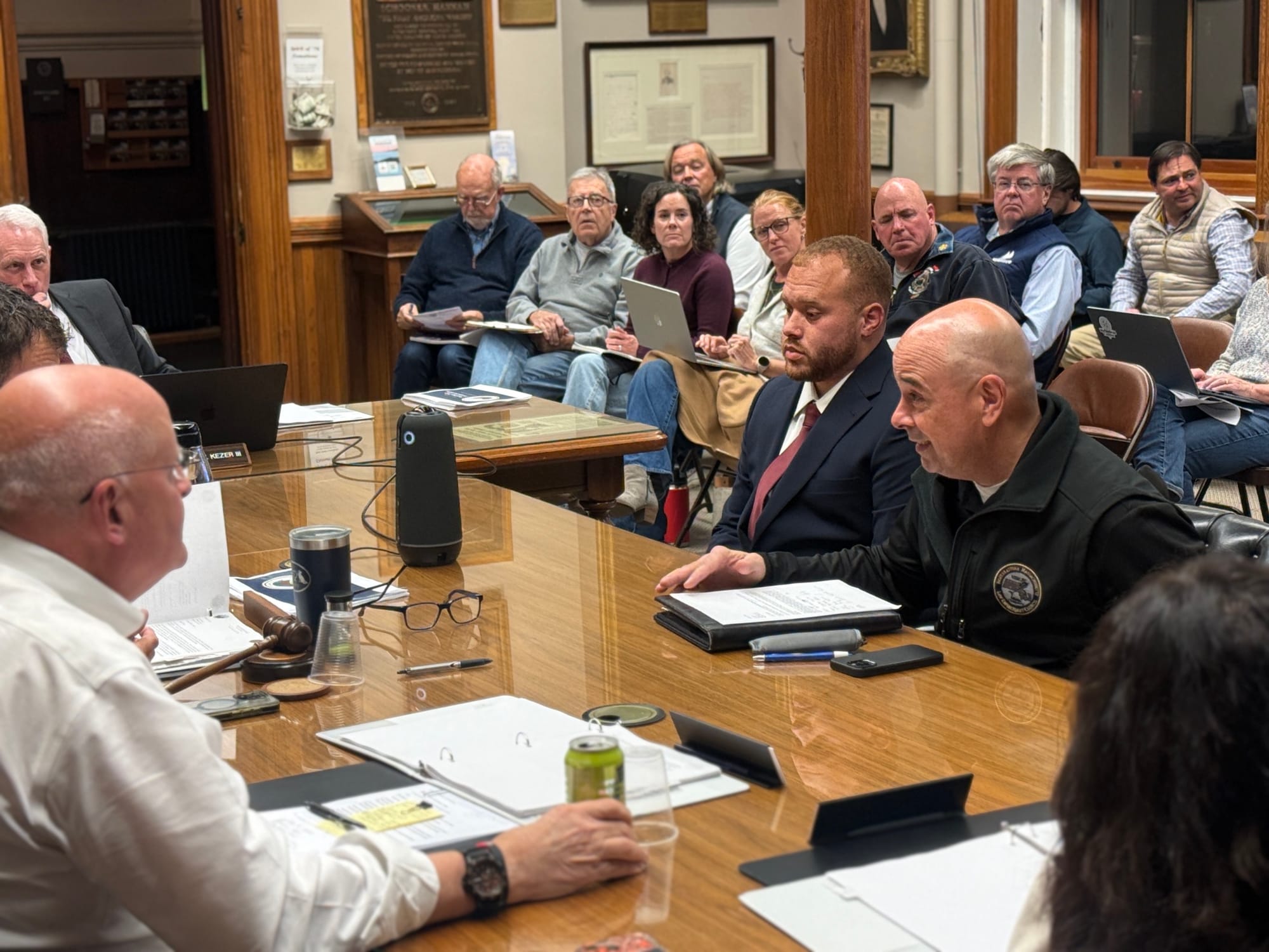 Two men sit side by side at a long wooden table during a meeting at Abbot Hall; the man on the right is speaking while an audience looks on.