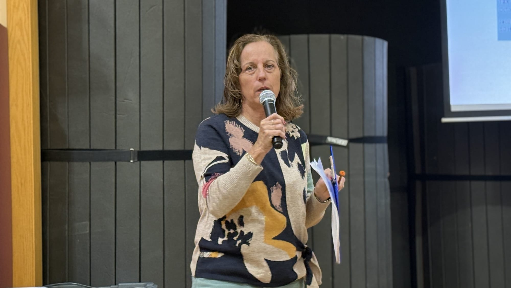 Jeanne Lambkin speaks into a microphone holding papers and a pen