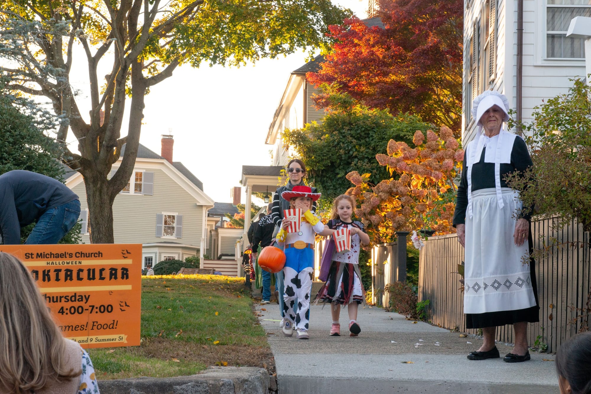 Children in costume walk along a sidewalk past a woman in a bonnet and apron
