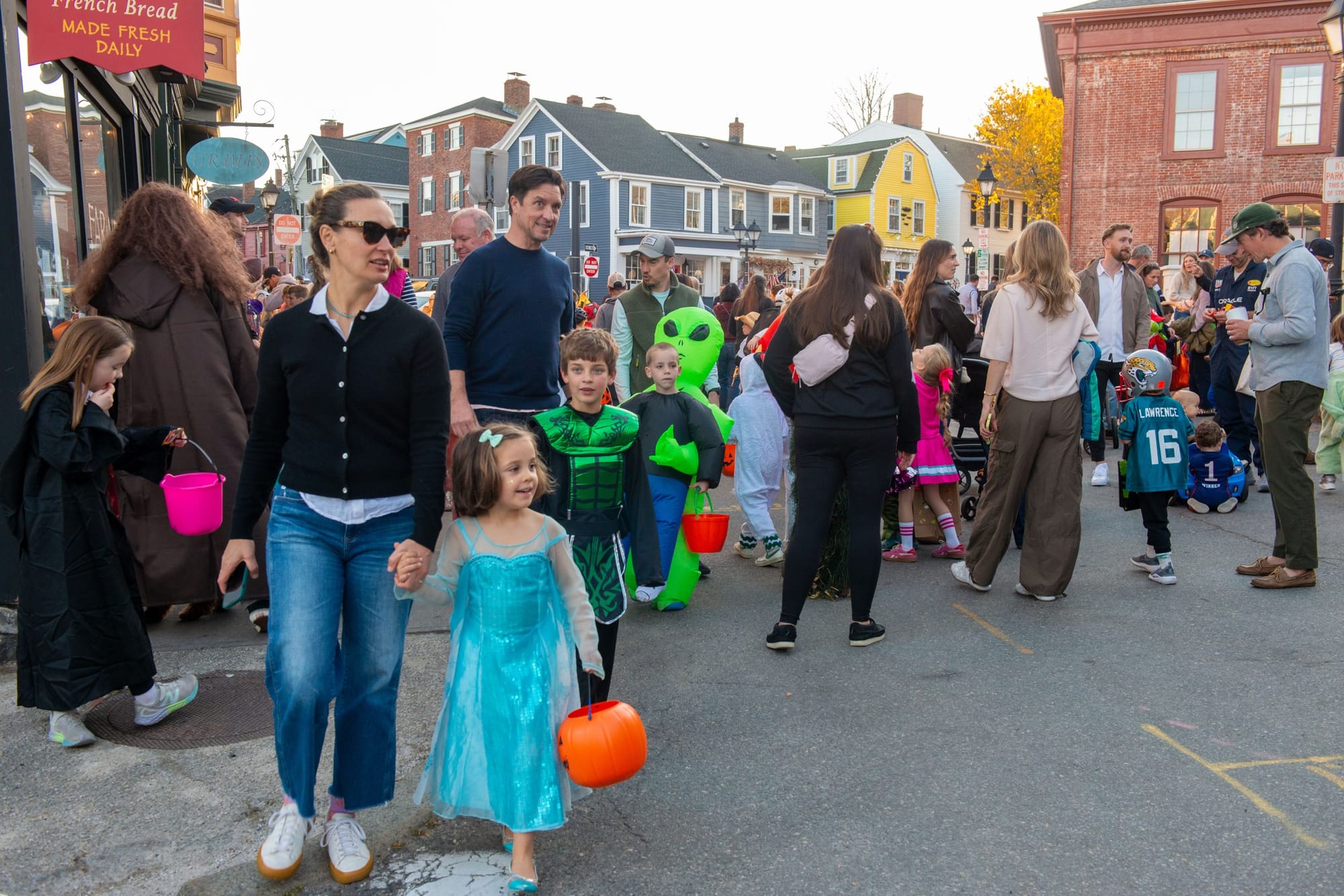 Girl in an Elsa costume with a pumpkin pail holds a woman's hand on a crowded street