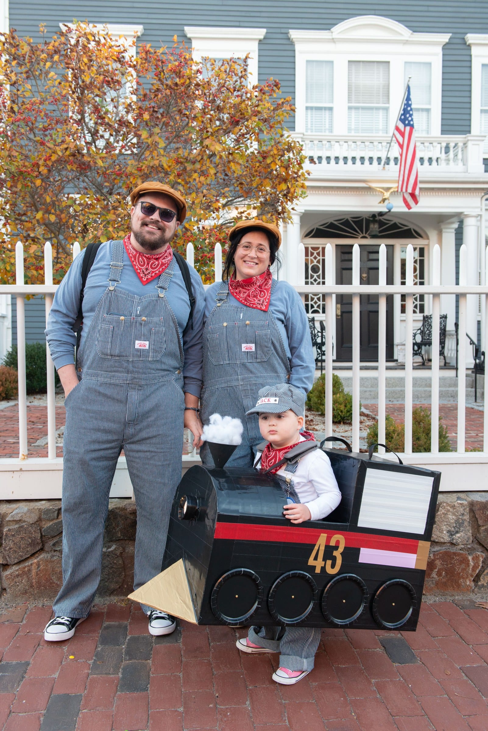 Two adults in striped overalls and bandanas stand beside a toddler in a cardboard No. 43 locomotive