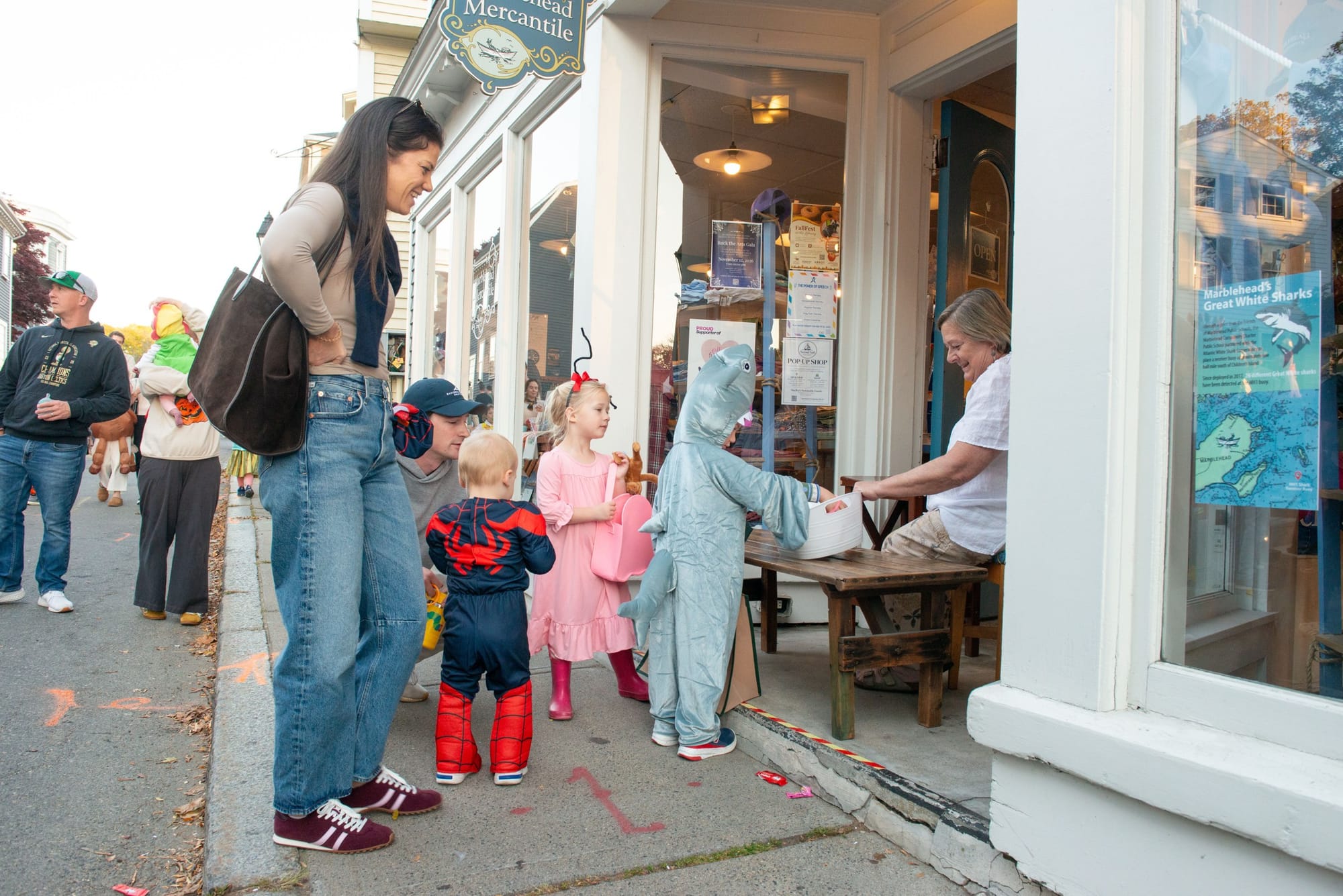 Child in a shark costume takes candy from a woman seated in a shop doorway