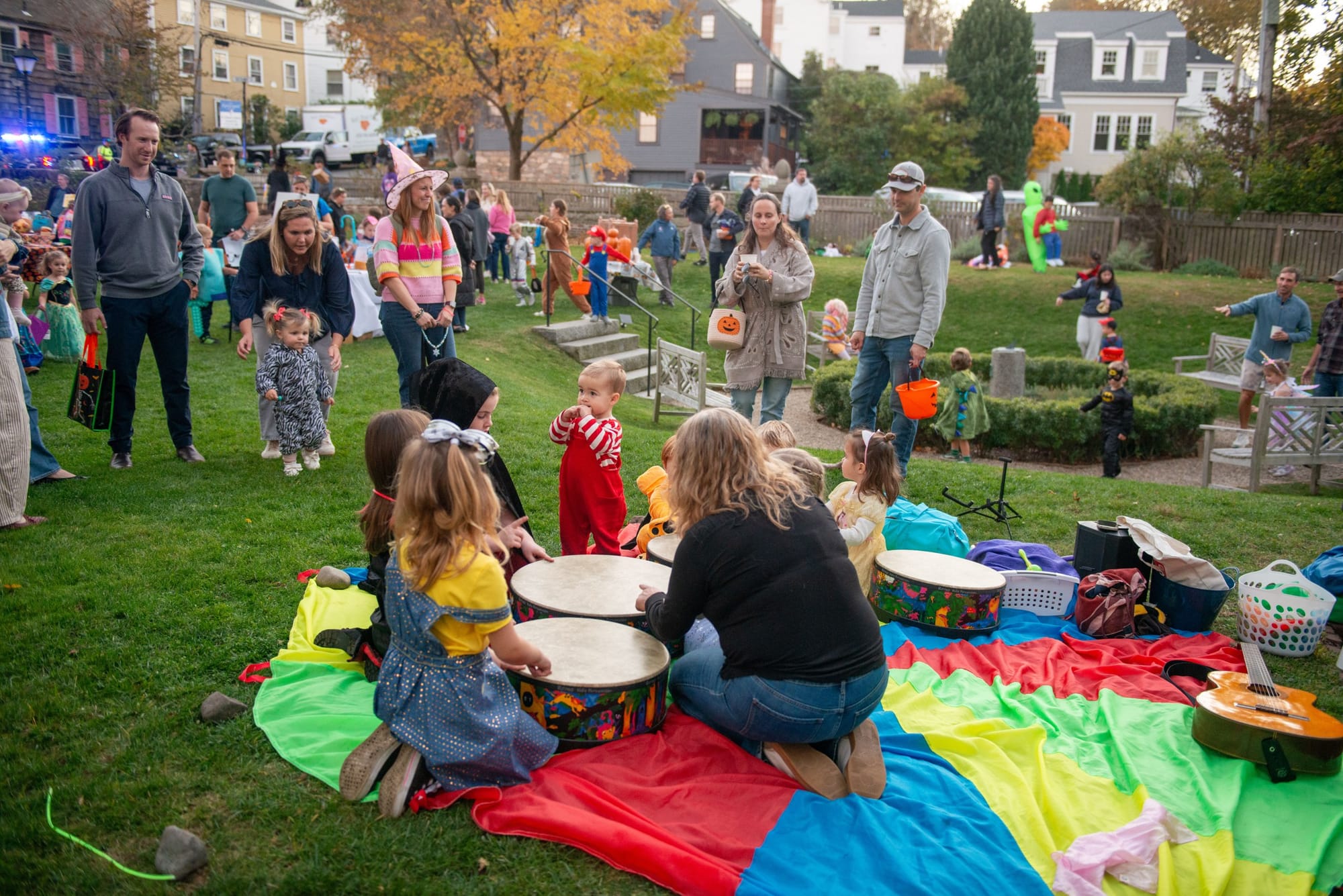 Children and adults play hand drums on a rainbow parachute on a lawn