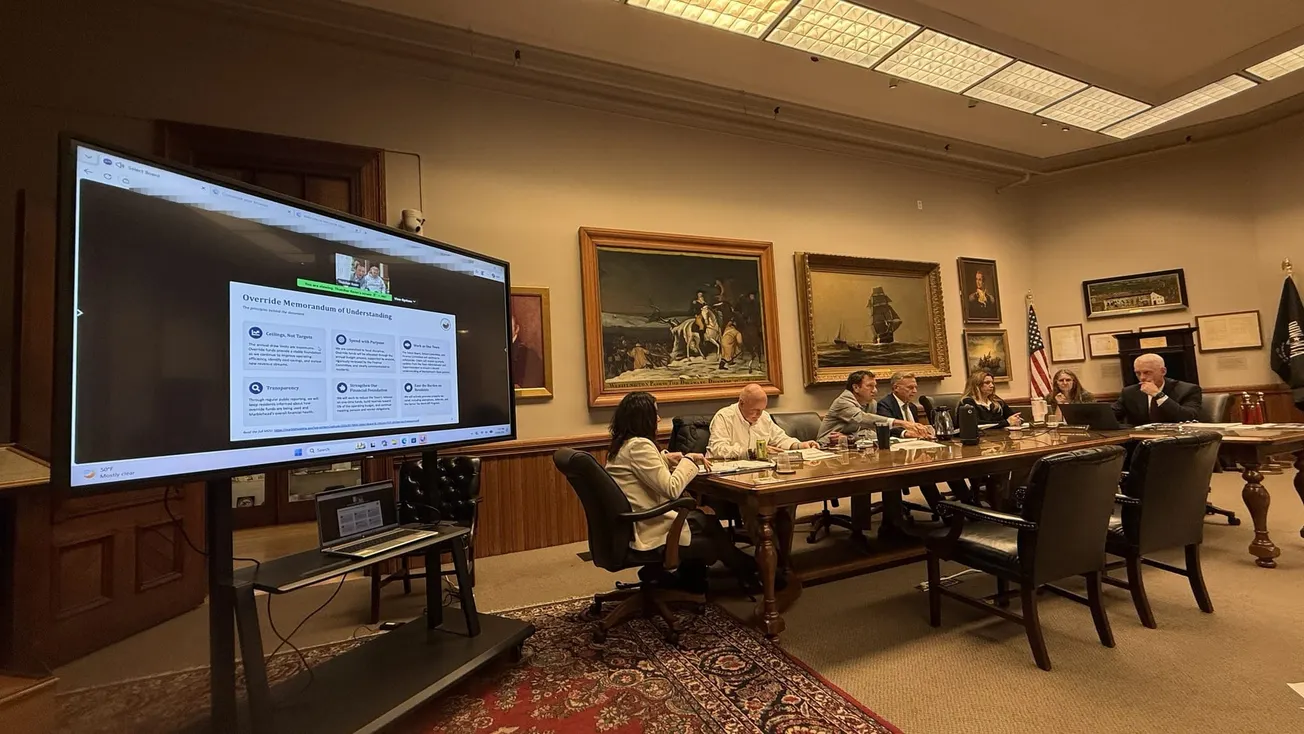 Town officials sit along a long wooden table beside a large screen displaying a slide titled Override Memorandum of Understanding.