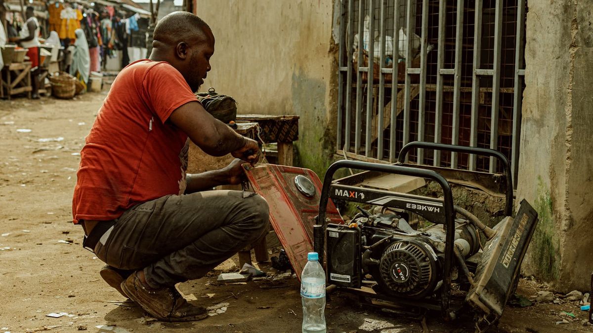 A man repairs a portable generator outside a building, with market stalls in the background.