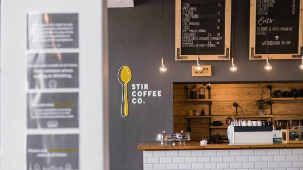 Handwritten drinks menus, a collection-point sign and a yellow spoon logo above the counter at Stir Coffee Co. in Gqeberha.