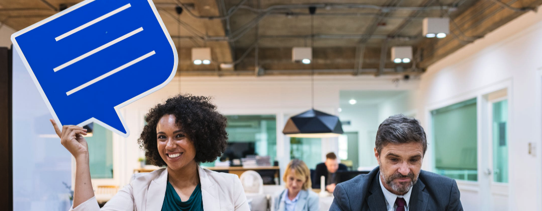A woman holds a large speech bubble sign while sitting next to a man in a business office.