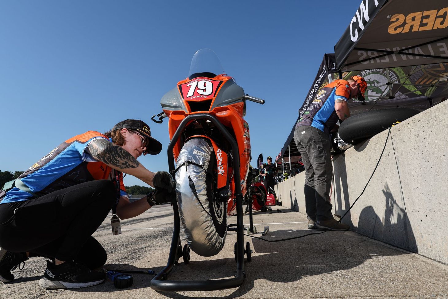 Sara is squatting down checking the front tire pressure on the orange and grey #79 CW Moto Racing Yamaha R1.