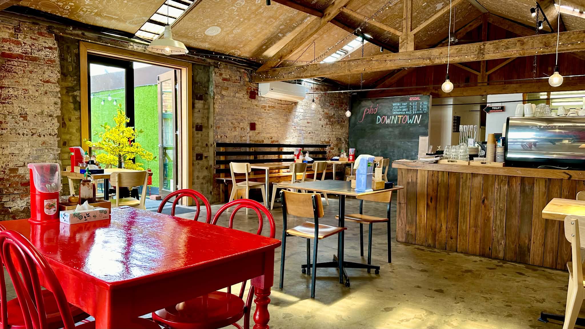 The interior of Pho Downtown, with exposed brick, timber beams, red tables and a handwritten menu on a dark wall.