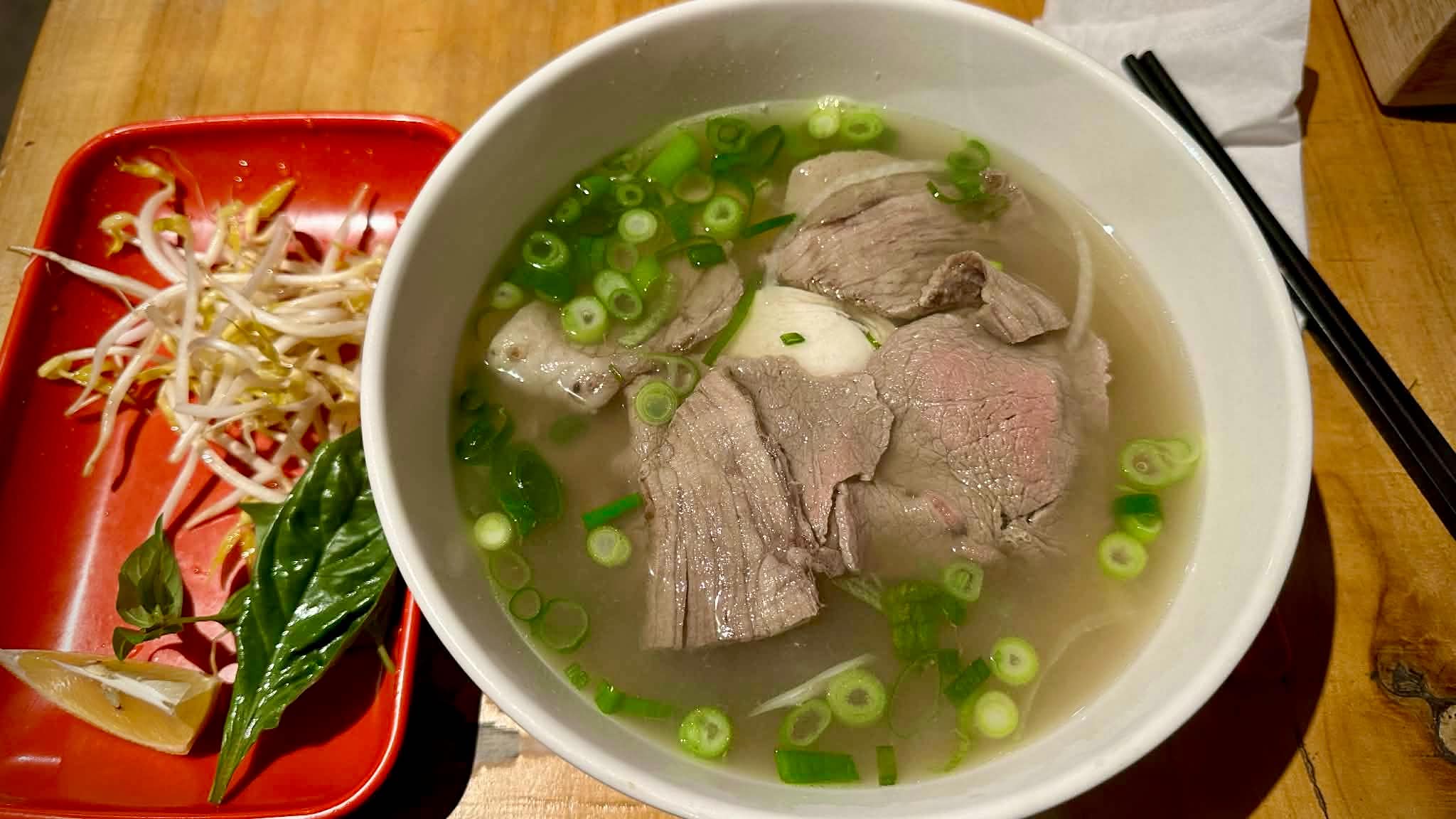 A white bowl of beef pho beside a red plate of bean shoots, basil and lemon on a timber table.