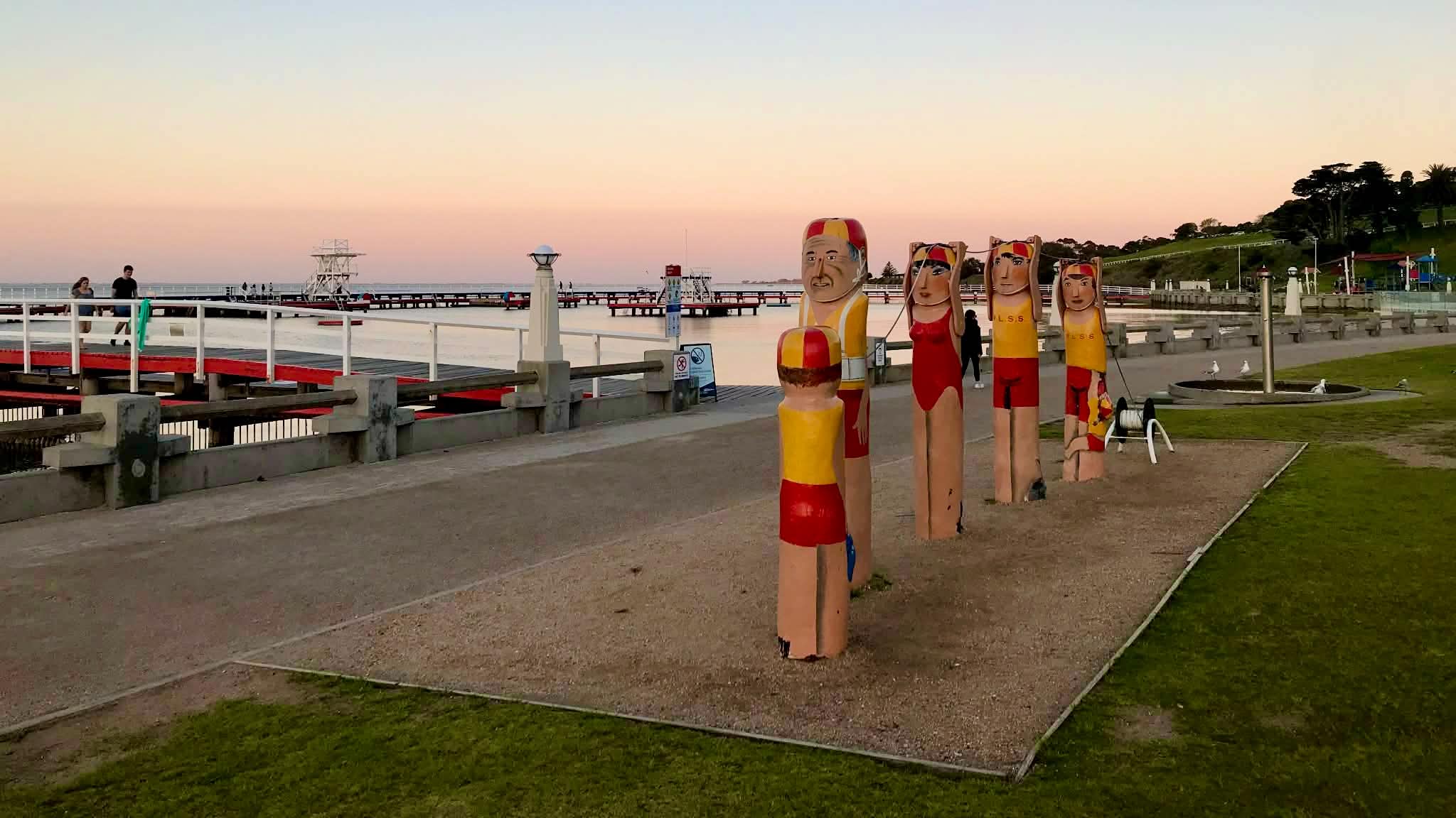 A row of red-and-yellow painted lifesaver bollards beside the Eastern Beach promenade, with the sea baths and Corio Bay beyond.