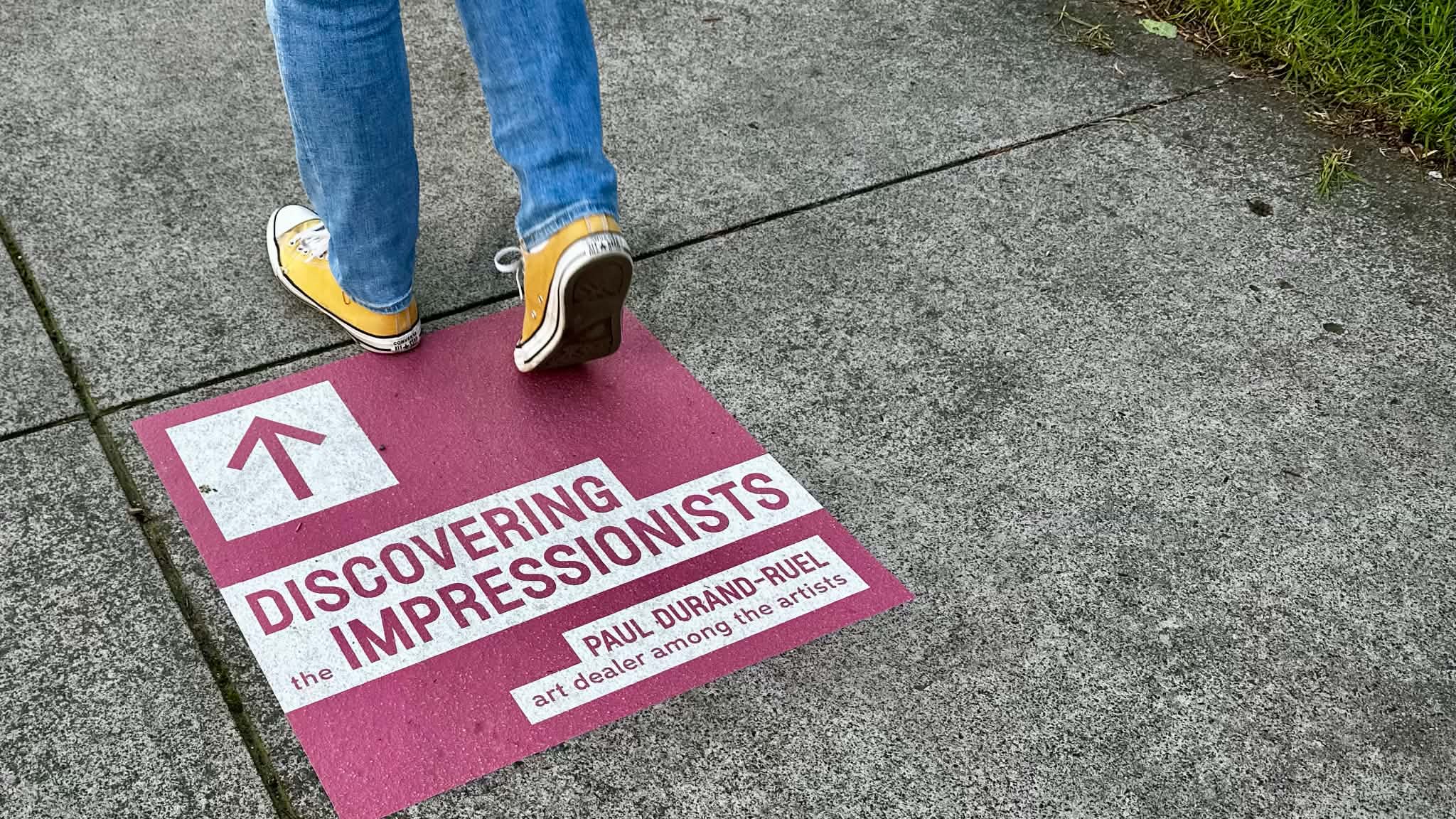 Jason in yellow shoes stepping across a magenta pavement sign pointing towards the Discovering the Impressionists exhibition.