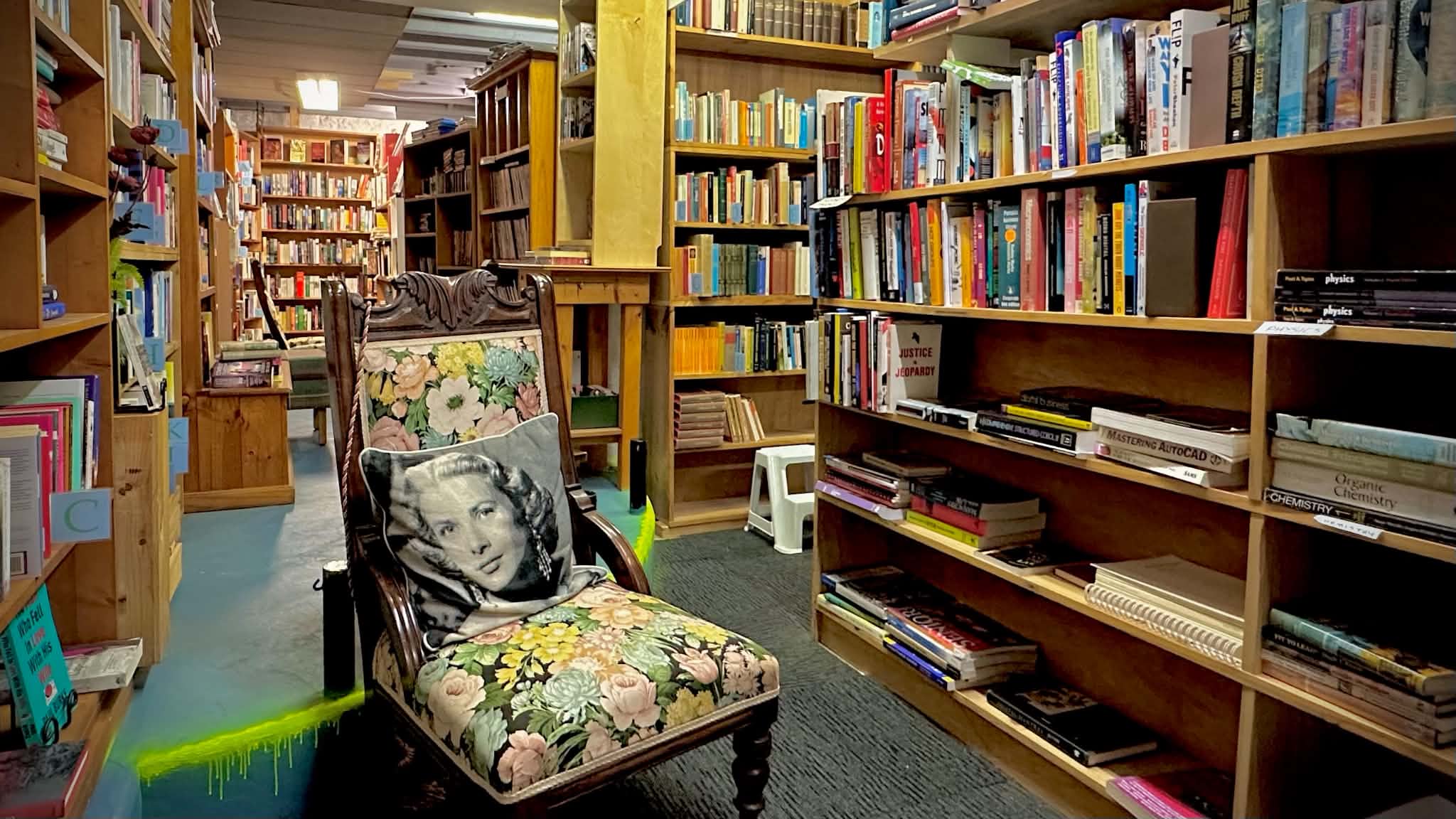 A floral armchair and portrait cushion sit among crowded shelves of secondhand books in the basement of Books Around.