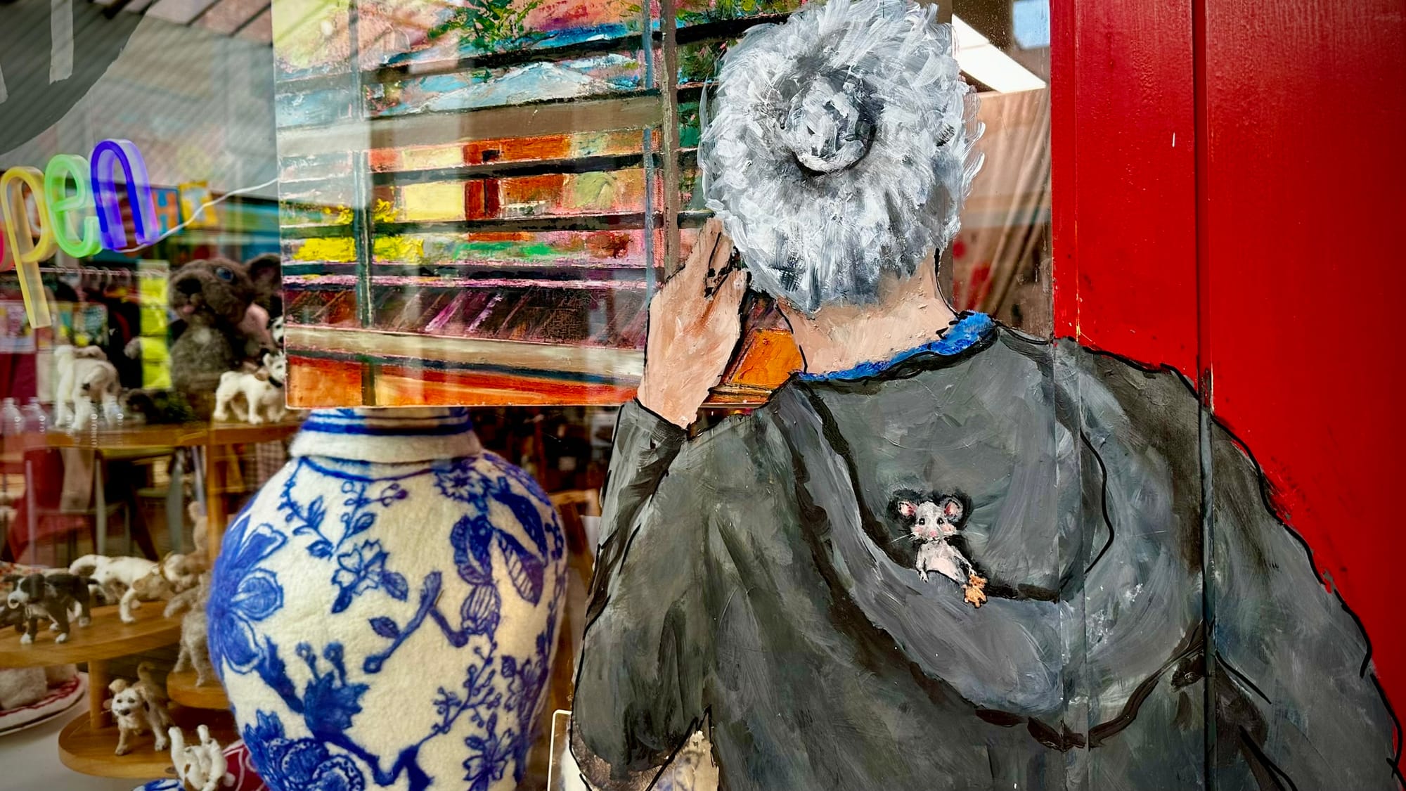 The window of Pip-Squeak Art Gallery and Studio, showing a painting of a woman, a blue-and-white vessel, felt animals and arcade reflections.