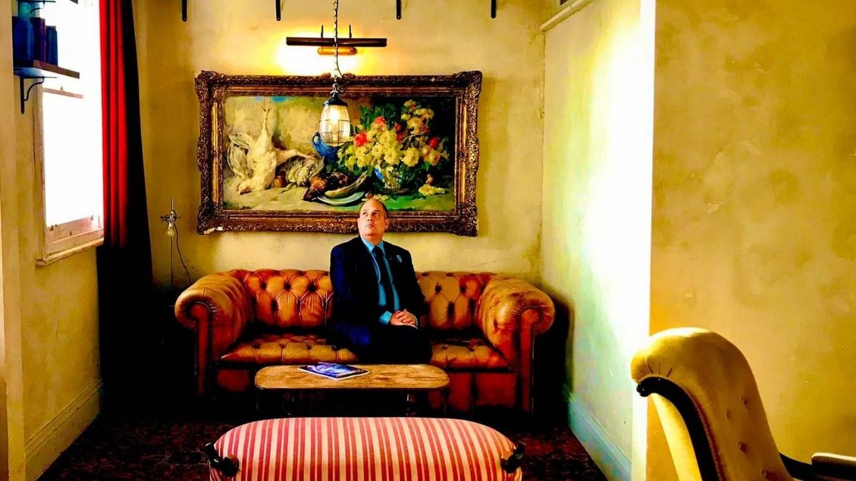 Graeme seated on a leather sofa beneath a large framed painting in a warmly lit interior.