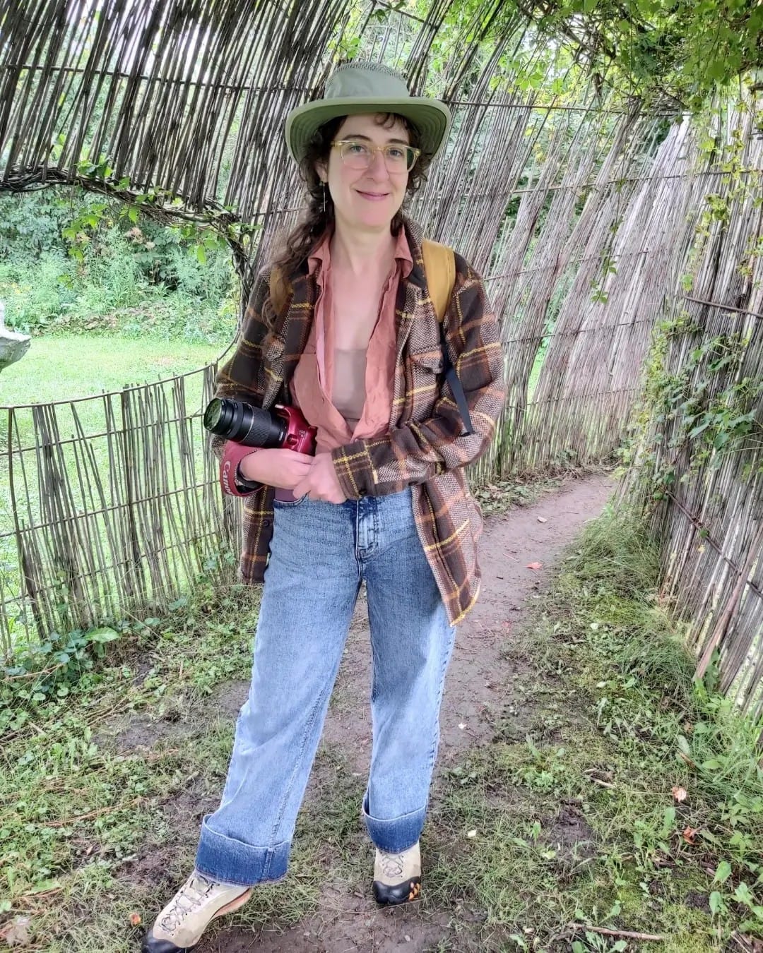 A photo of Idzie, a white person with long brown hair wearing a Tilley hat and plaid jacket, with a red DSLR camera cradled in her arms. She's standing in a tunnel made of willow sticks and smiling at the photographer.