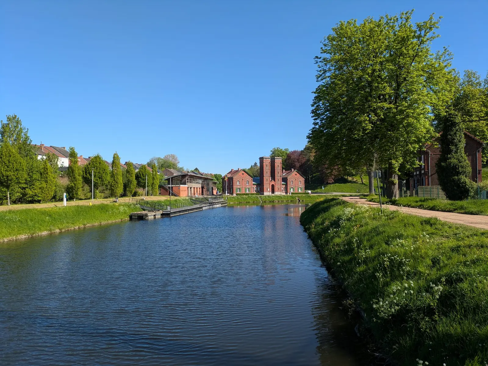 Canal paisible bordé d’arbres et de maisons en briques sous un ciel bleu, avec une écluse rouge au loin.