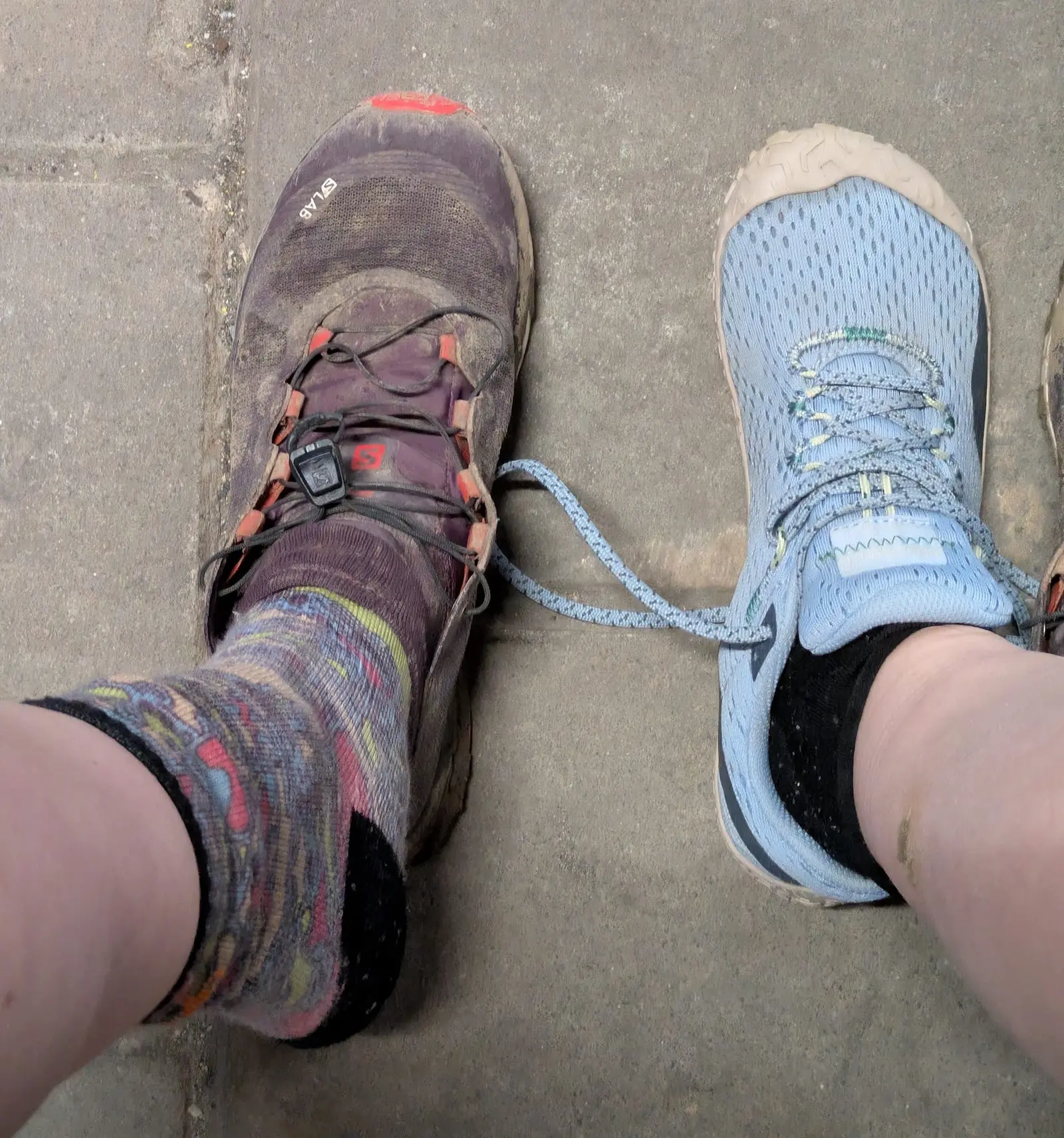 Deux chaussures de randonnée sales, lacets emmêlés, sur un sol en pierre, avec chaussettes colorées et bleue.