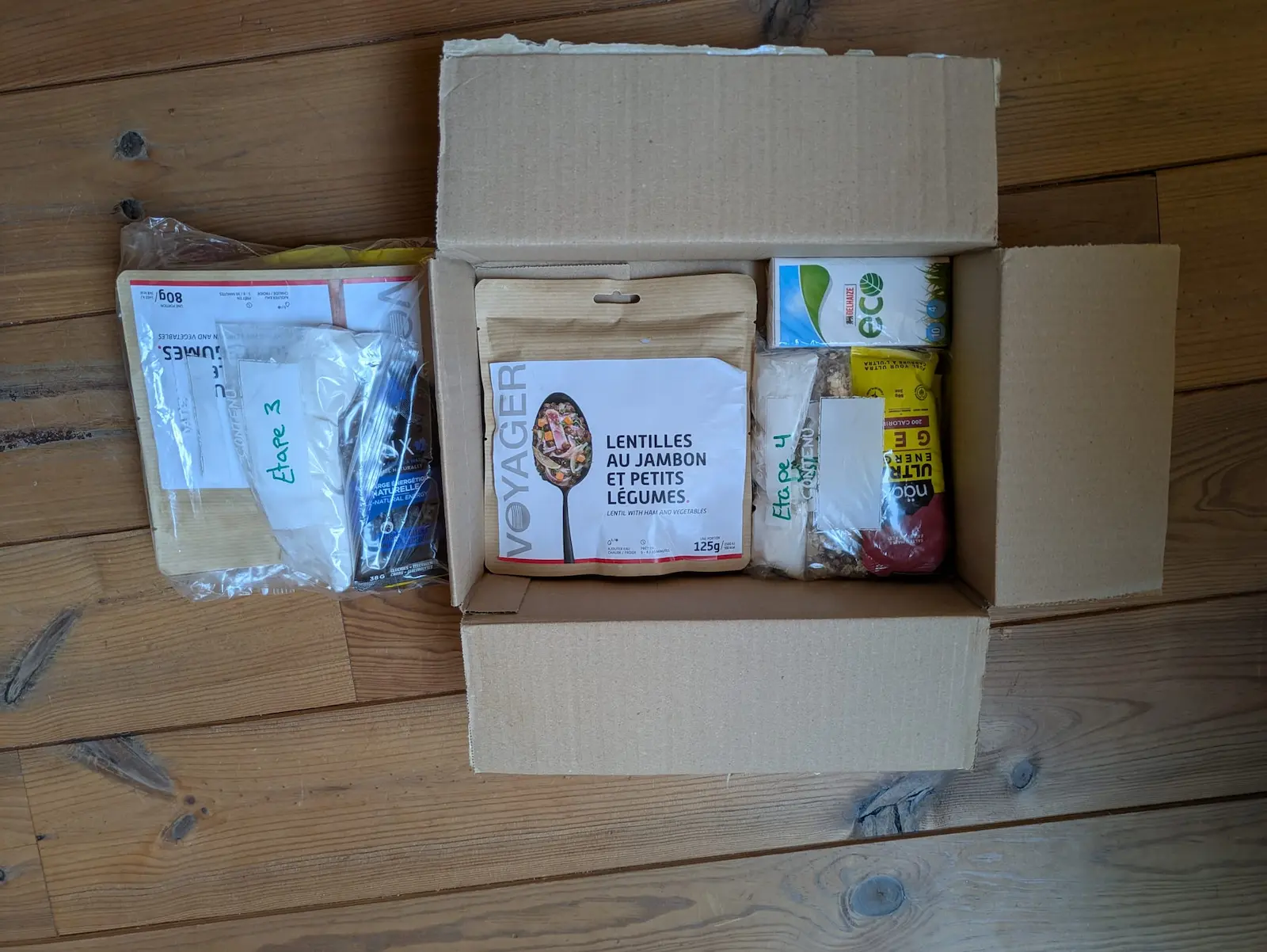 Carton ouvert sur parquet, rempli de repas et sachets alimentaires étiquetés, dont LENTILLES AU JAMBON ET PETITS LÉGUMES.