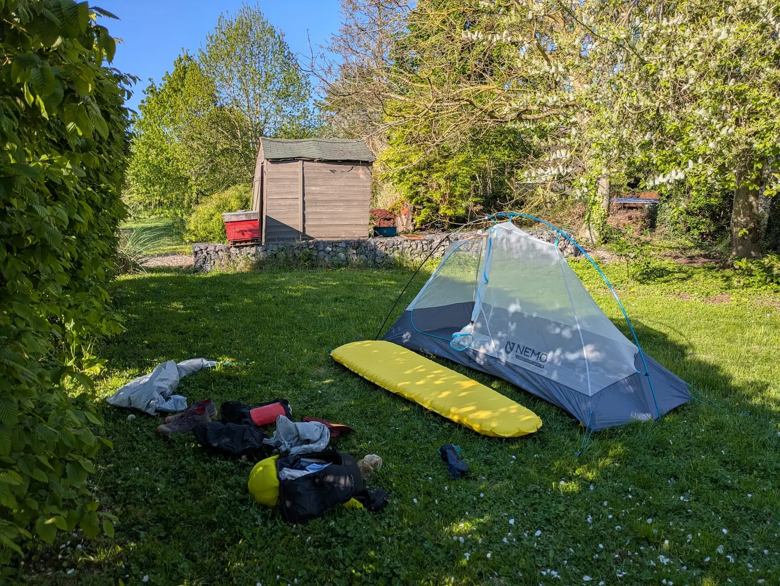 Tente transparente avec tapis jaune sur l'herbe verte, entourée de sacs et chaussures. Abri en bois en arrière-plan, atmosphère paisible.
