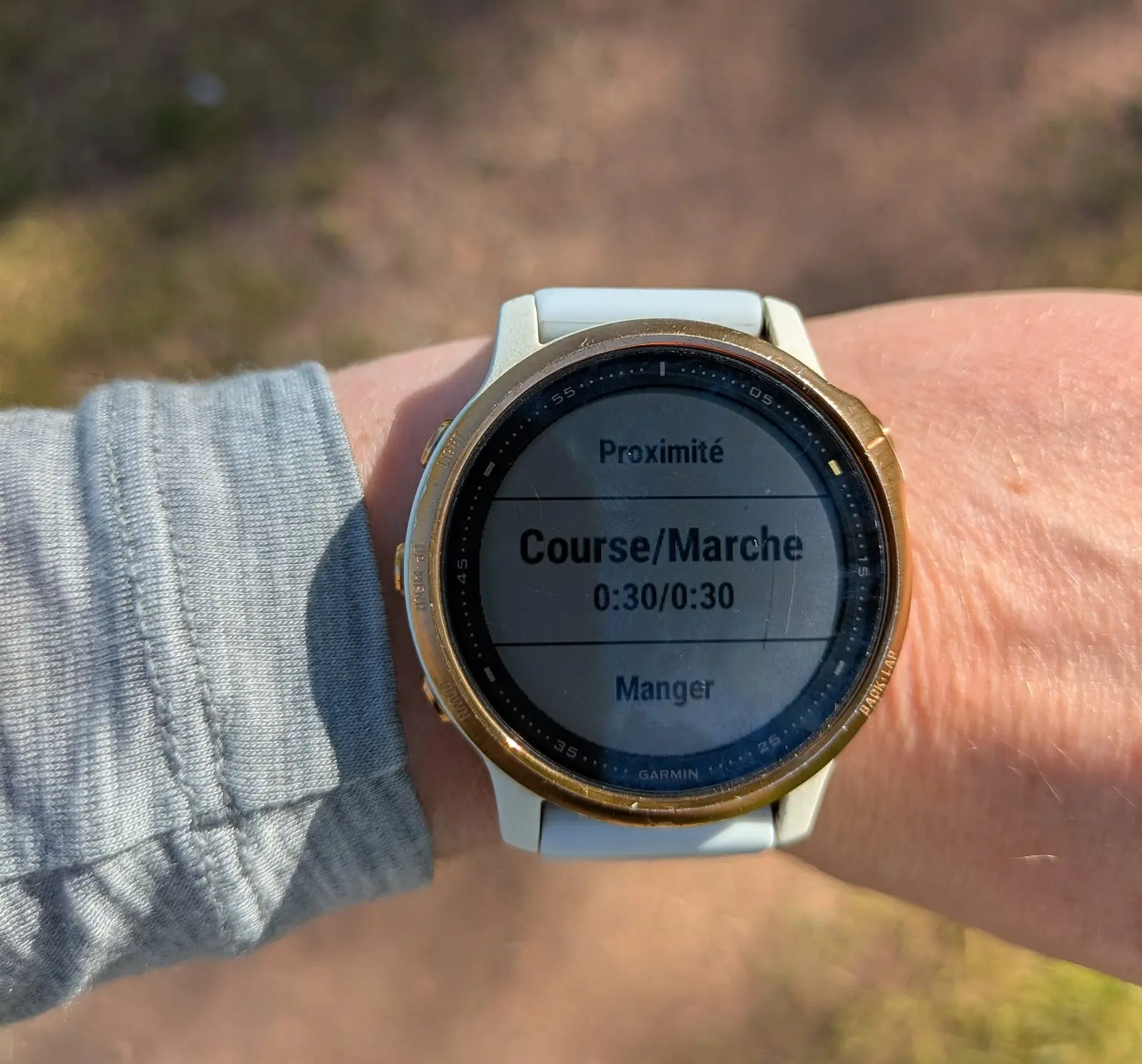 Montre Garmin au poignet affichant Course/Marche 0:30/0:30, sur fond de sentier flou en plein soleil.