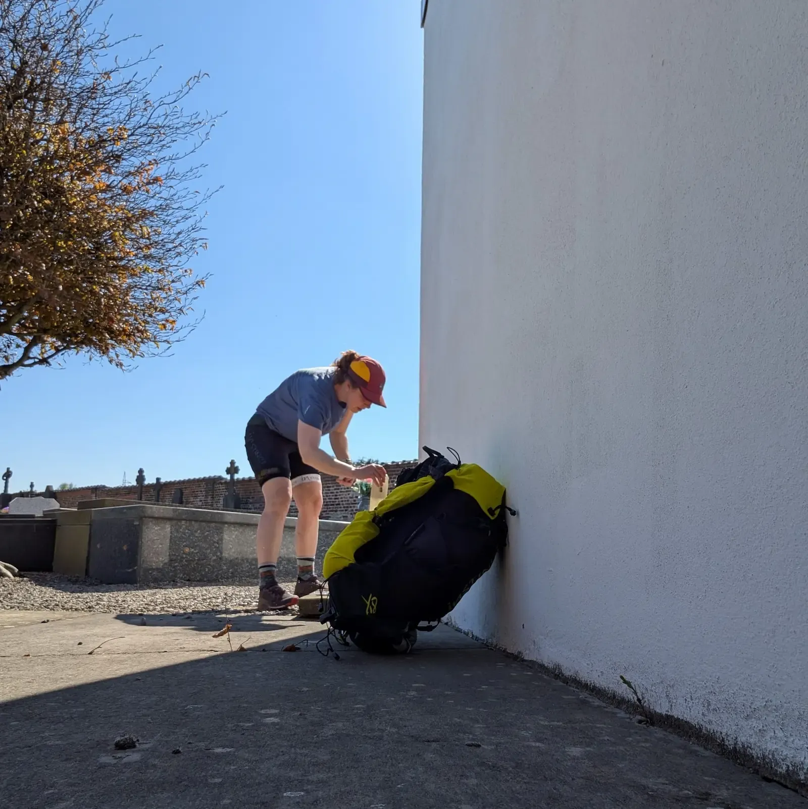 Personne en short et casquette ajuste un sac à dos jaune et noir contre un mur blanc, sous un ciel bleu.