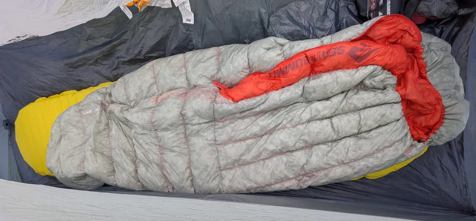 Sac de couchage gris, jaune et rouge dans une tente, sur tapis sombre; étiquettes visibles, ambiance de bivouac.