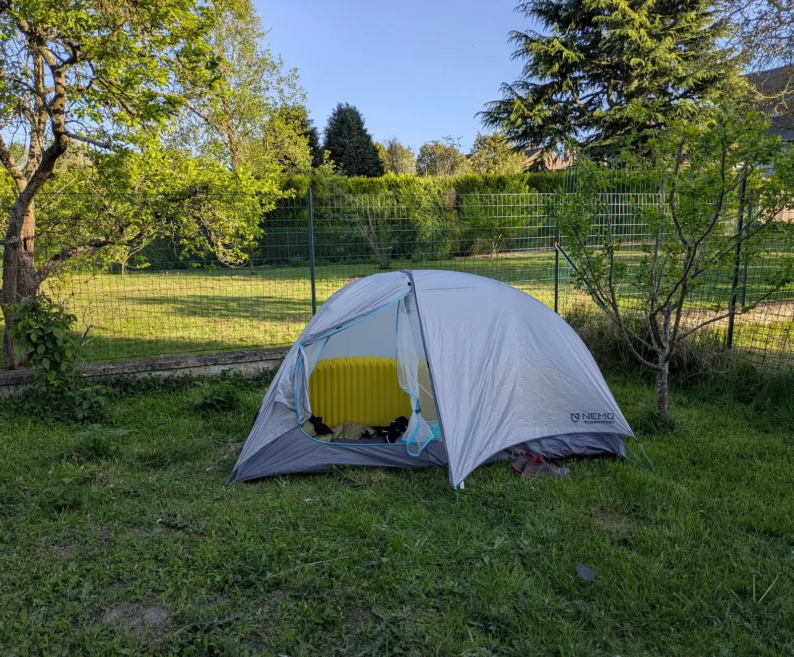 Tente grise sur pelouse verte, entourée d'arbres et clôture. Un matelas jaune à l'intérieur. Temps clair, ambiance paisible.