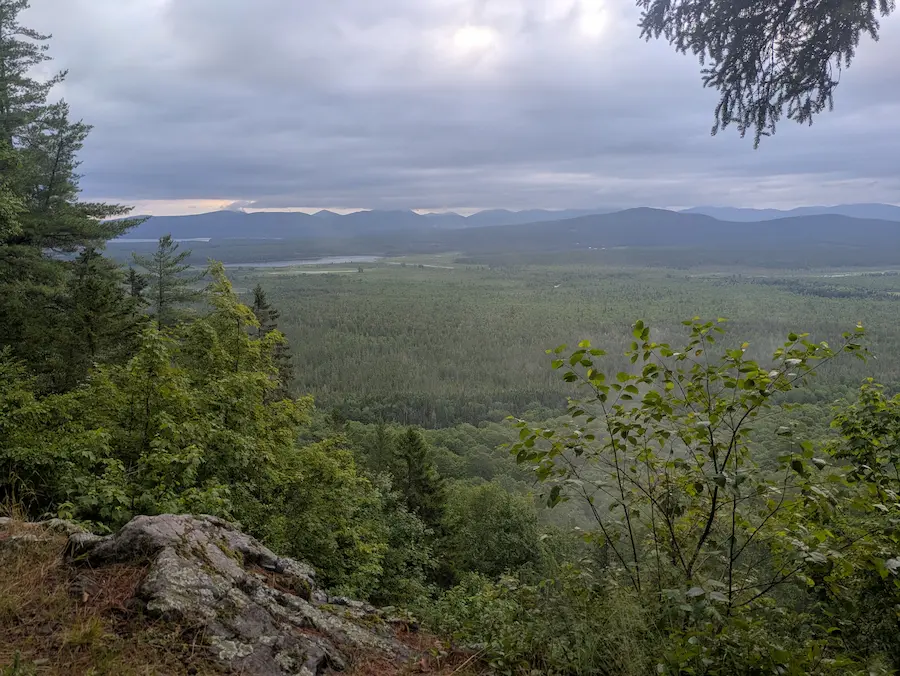 Paysage montagneux sous un ciel nuageux, forêt dense au premier plan, chaînes de montagnes à l'horizon, ambiance paisible et verdoyante.