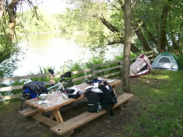 Vélos et équipement de camping sur une table en bois près d'une rivière. Tentes installées, ambiance paisible sous arbres verts.