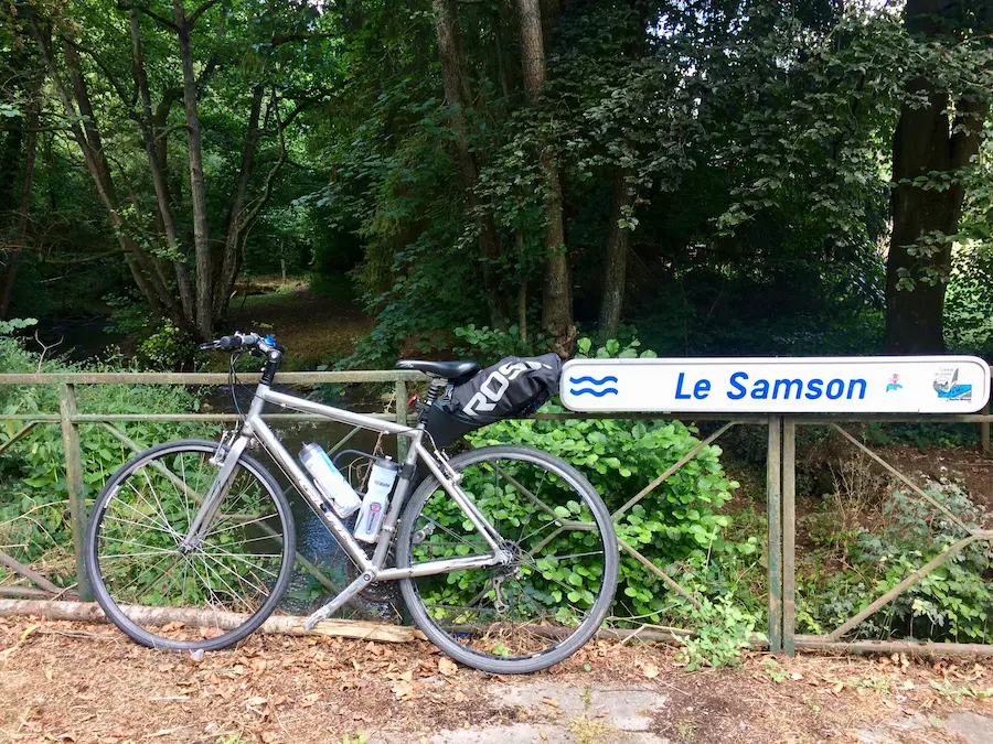 Vélo argenté appuyé contre une barrière devant un panneau "Le Samson", forêt luxuriante en arrière-plan, ambiance paisible.