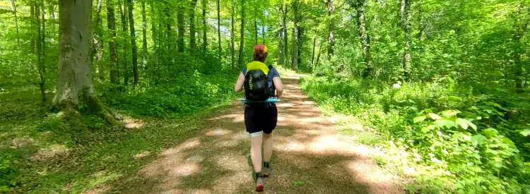 Personne vue de dos courant sur un chemin forestier ensoleillé, sac à dos noir et jaune, ambiance paisible.