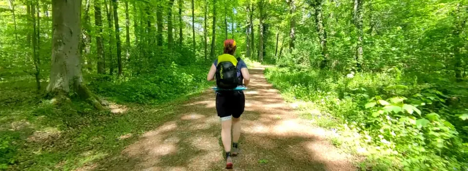 Personne vue de dos courant sur un chemin forestier ensoleillé, sac à dos noir et jaune, ambiance paisible.