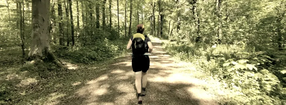 Personne vue de dos courant sur un chemin forestier ensoleillé, sac à dos noir et jaune, ambiance paisible.