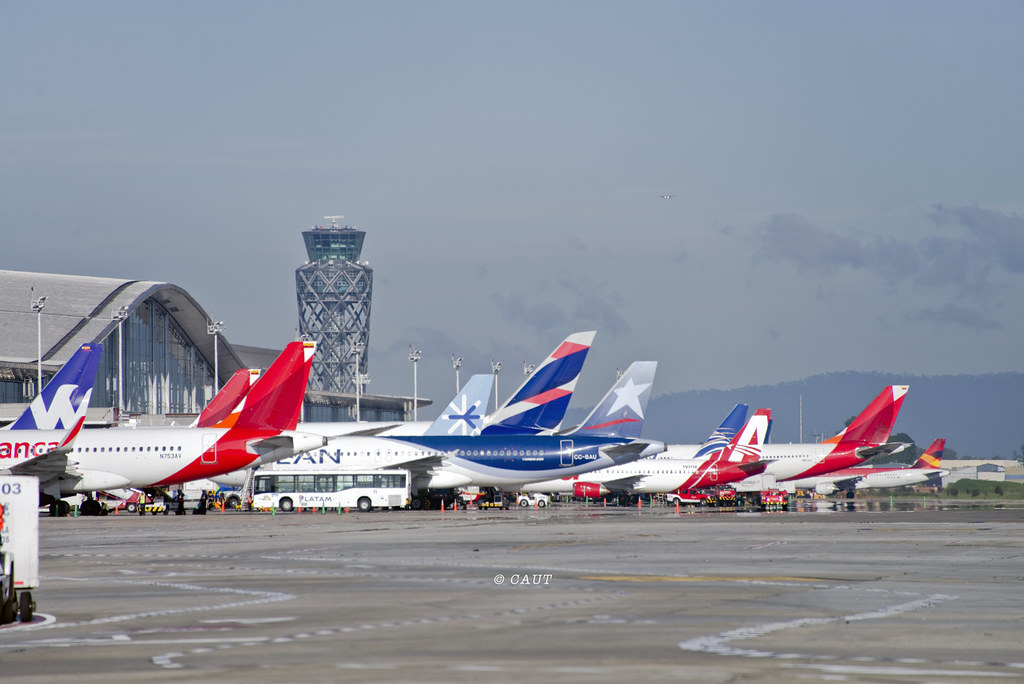 Row of commercial aircraft from various airlines parked at an airport terminal, with their colorful tails lined up in front of the control tower.