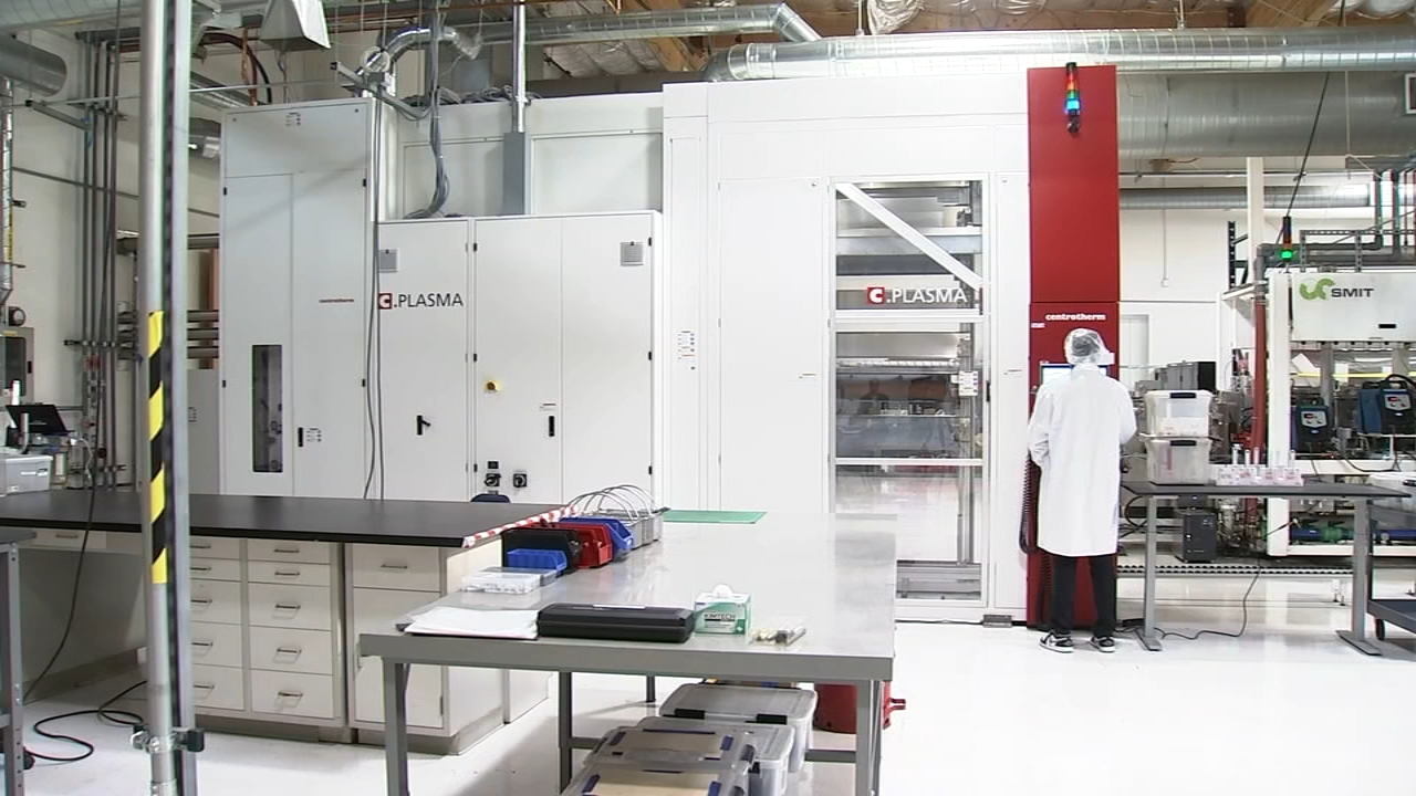 Person in a white lab coat working with industrial plasma equipment inside a clean manufacturing facility with various machines and workstations
