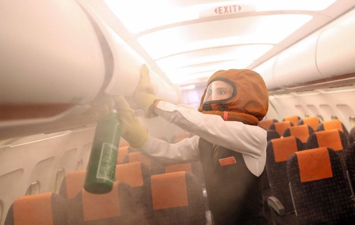 A flight attendant wearing protective headgear and gloves uses a fire extinguisher to put out smoke inside an airplane cabin, with orange passenger seats and an exit sign visible above.