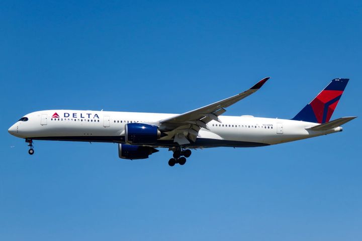 A Delta Air Lines Airbus A350 aircraft in flight against a clear blue sky, with landing gear extended and the airline’s red-and-blue tail logo visible.