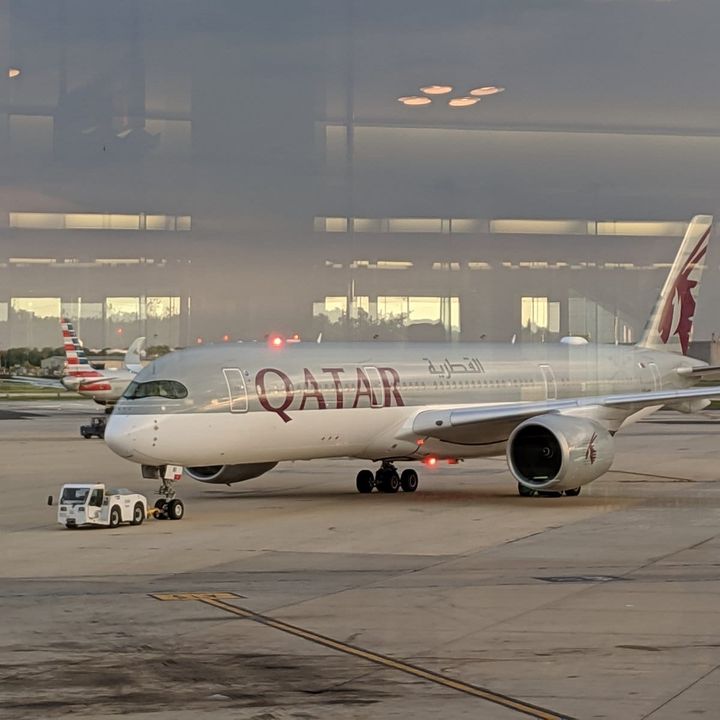Qatar Airways aircraft on the tarmac at an airport, with an American Airlines plane visible in the background.