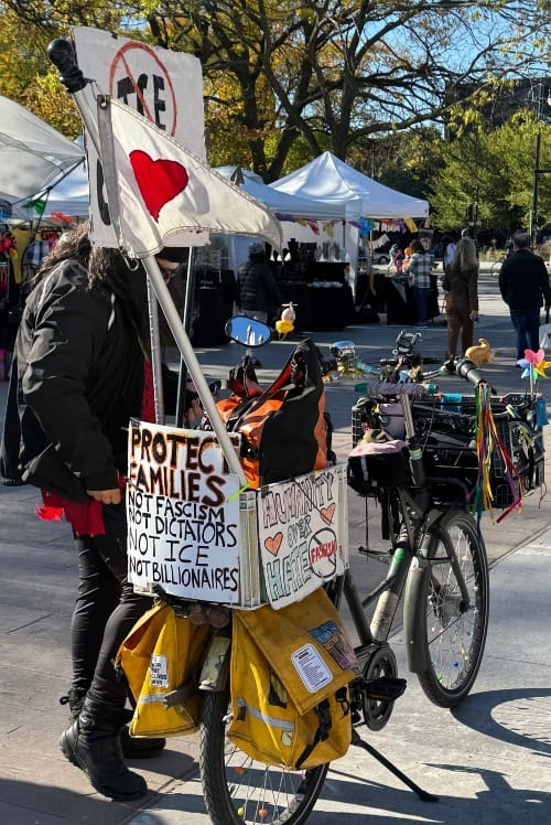 Photograph of a person in black winter clothing, standing beside a yellow bicycle with a colorful cargo trailer. The trailer has protest signs, including "PROTECT FAMILIES, NOT FASCISM, NOT DICTATORS, NOT ICE, NOT BILLIONAIRES." Background features white vendor tents, trees with yellow leaves, and a sunny Lincoln Square scene.